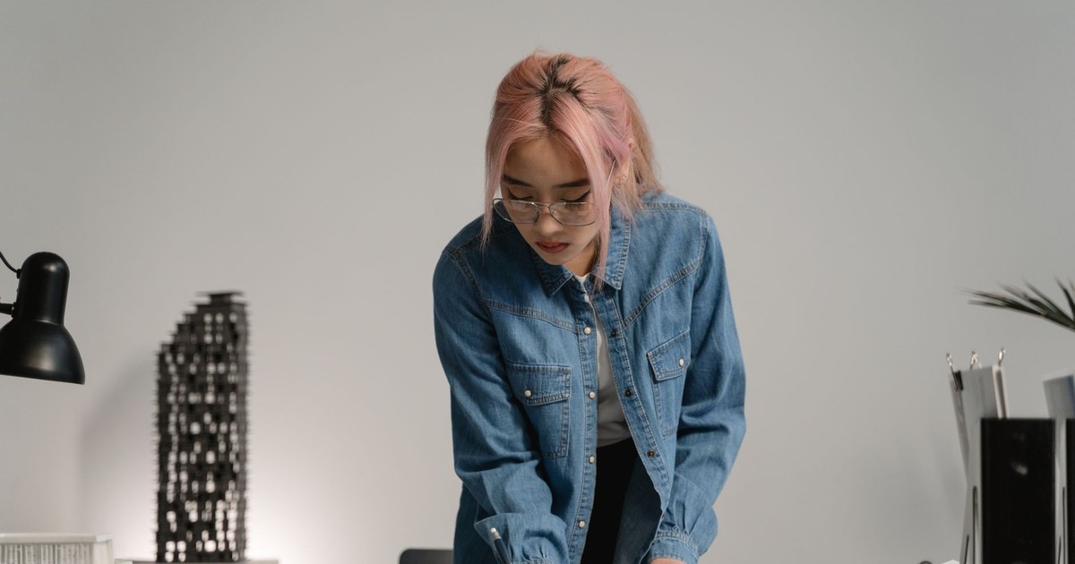 Asian woman with pink hair in a denim jacket focusing on work in an indoor setting.