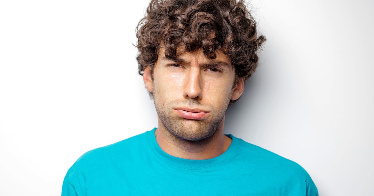 Portrait of a curly-haired man in a blue shirt with a grumpy expression against a white background.