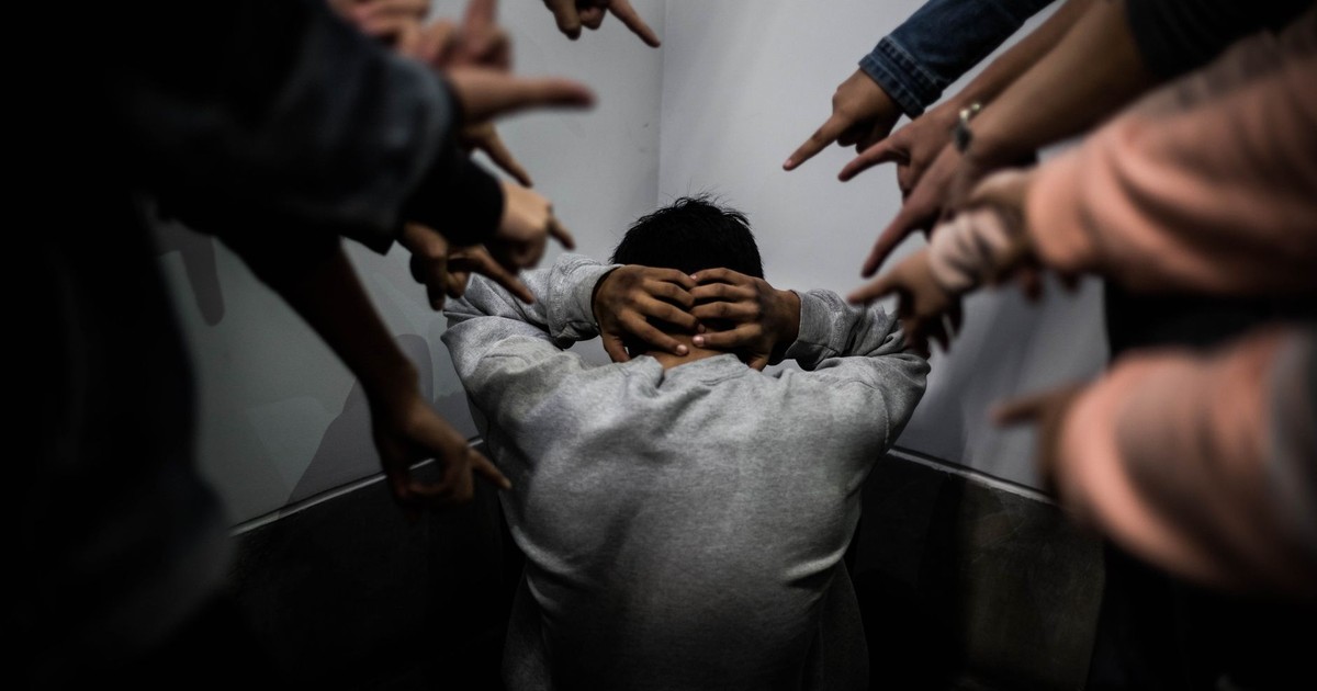 A person crouches in a corner surrounded by pointing fingers, illustrating social pressure and bullying.