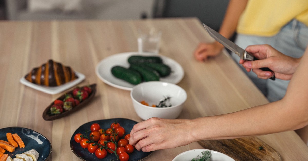 Hands slicing and arranging fresh vegetables and fruits on wooden table indoors.