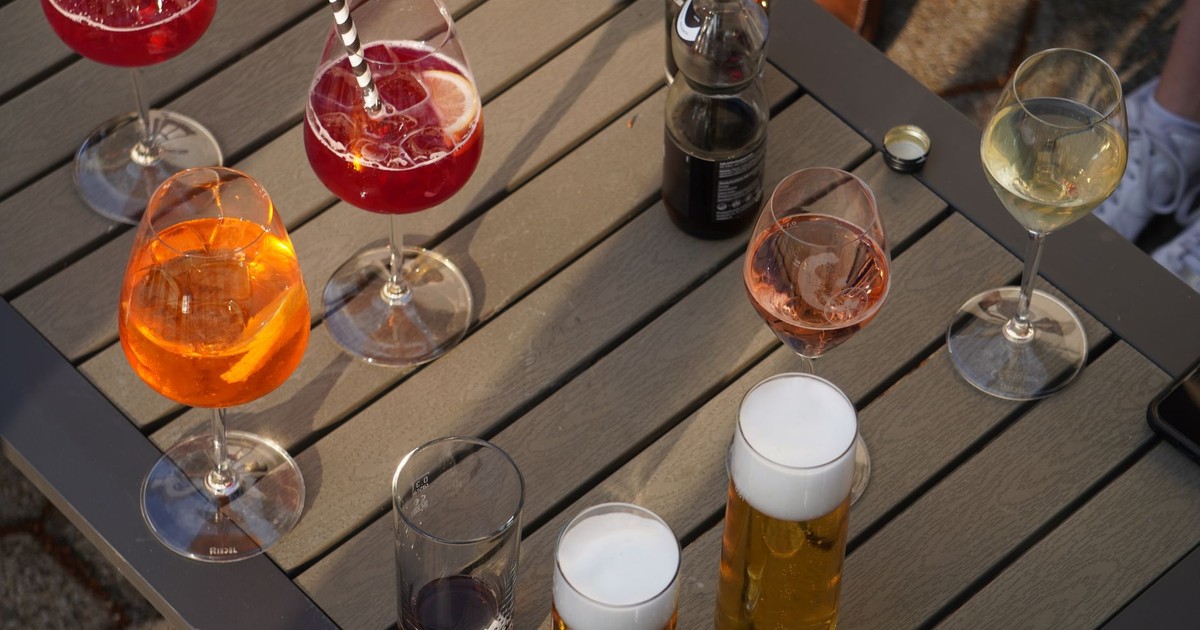 A variety of colorful drinks including cocktails, wine, and beer are arranged on a wooden table outdoors.
