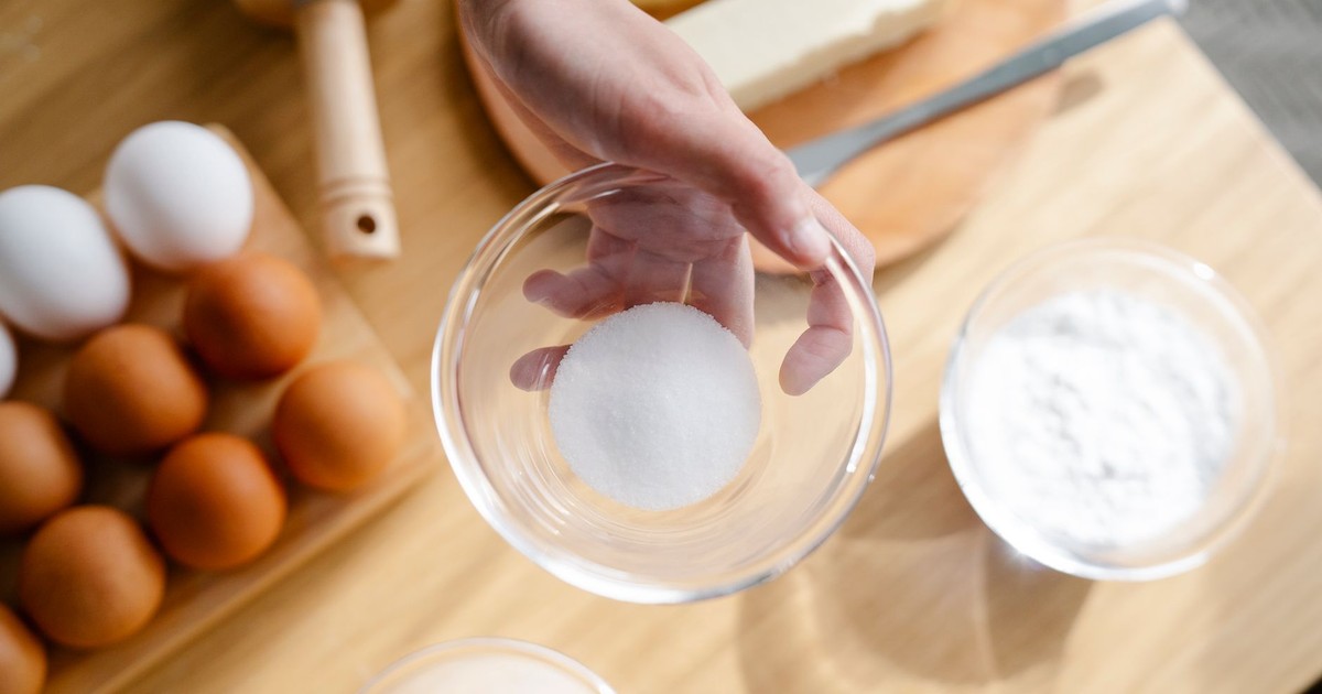 Close-up of hand holding sugar in glass bowl with baking ingredients like eggs and flour.