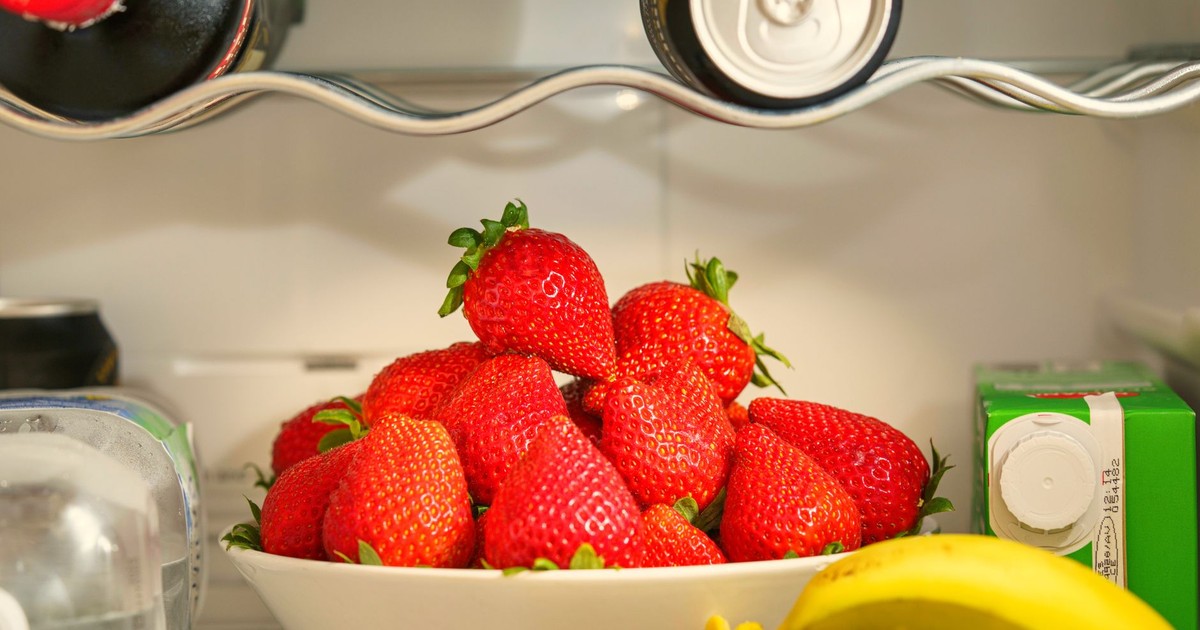 A bowl of ripe strawberries in a refrigerator, surrounded by drinks and bananas, showcasing vibrant freshness.