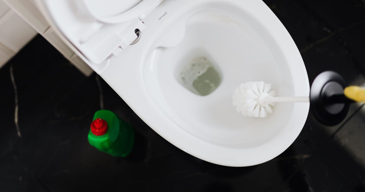 From above crop anonymous person in yellow gloves cleaning toilet bowl with toilet brush and liquid detergent in green plastic bottle