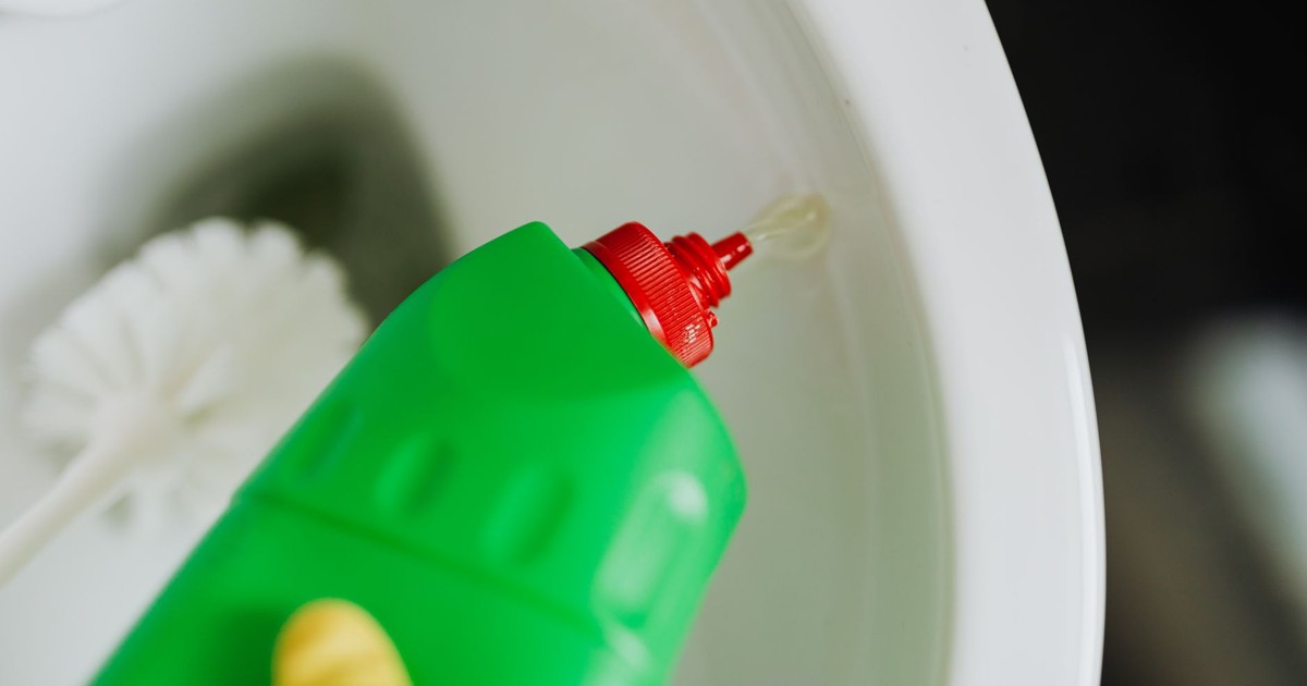 Top view of crop faceless person in yellow rubber gloves cleaning toilet with brush and pouring liquid disinfecting cleaner in toilet bowl