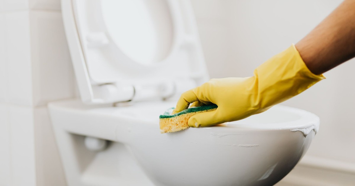 Crop anonymous person in yellow gloves cleaning white toilet rim with detergent and sponge in bathroom