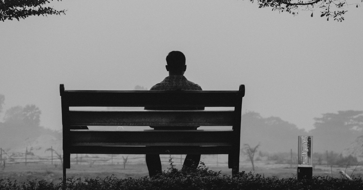 A solitary man sits on a bench in Dhaka, Bangladesh, silhouetted against a foggy, grayscale landscape.