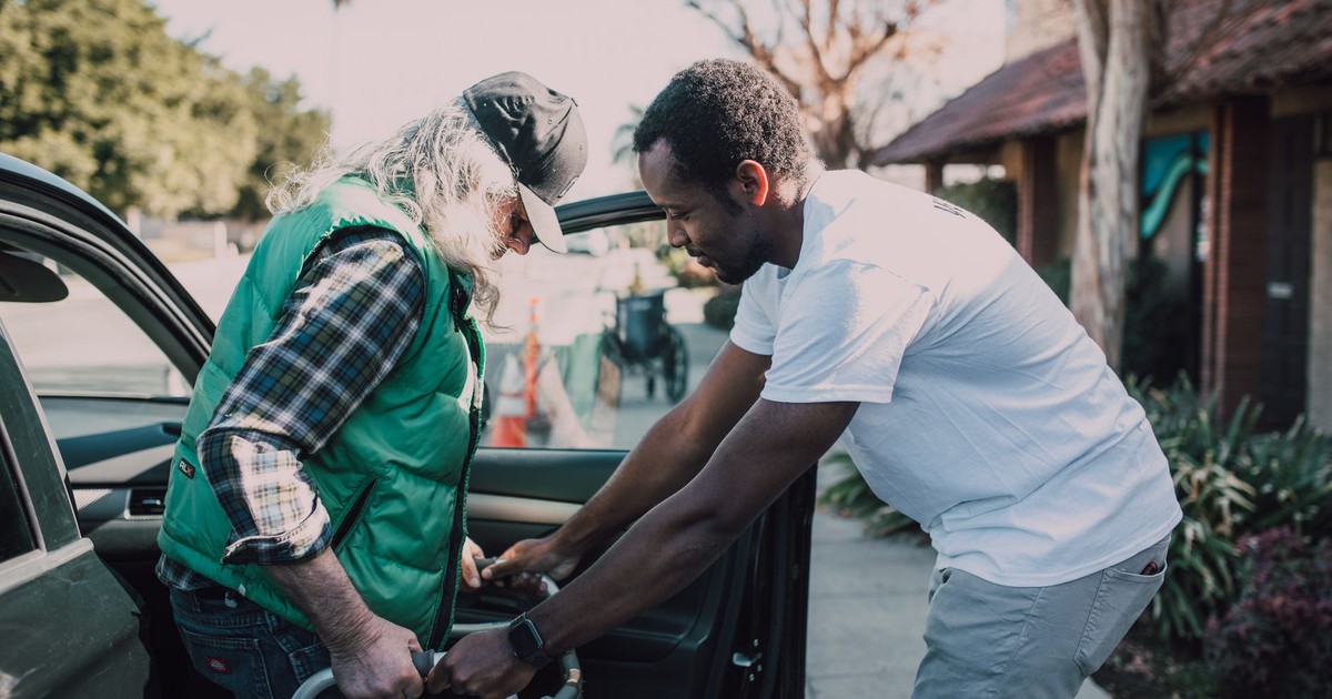 Young volunteer helping senior man with walker near a parked car, symbolizing care and support.