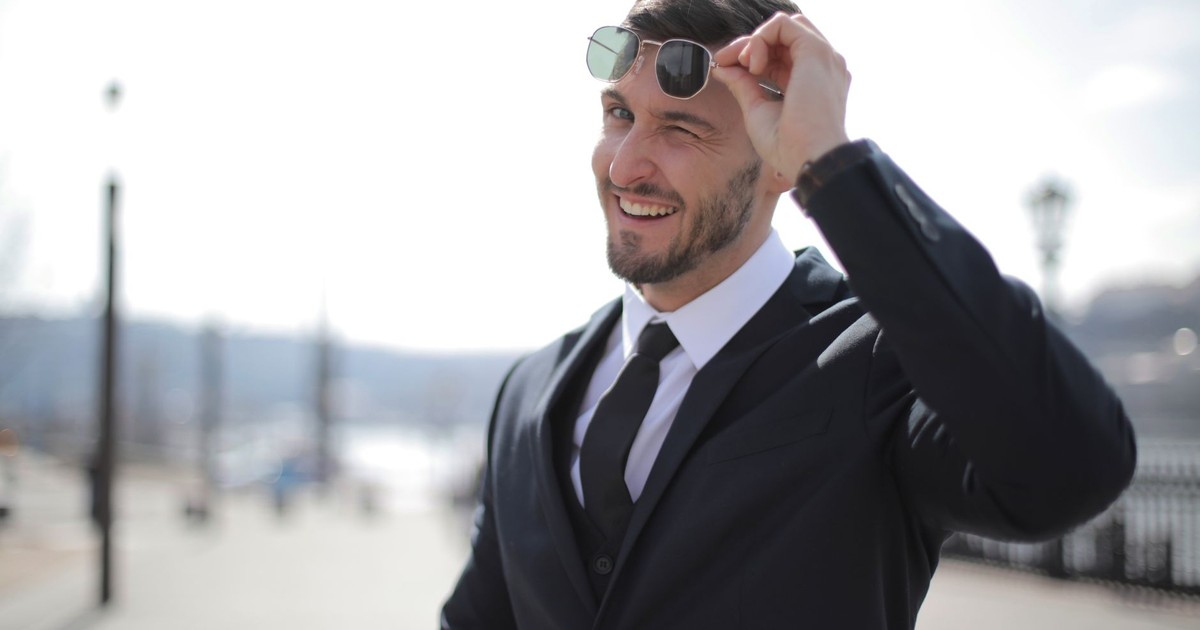 A charming man in a suit winks while holding sunglasses outdoors, exuding a confident and friendly demeanor.