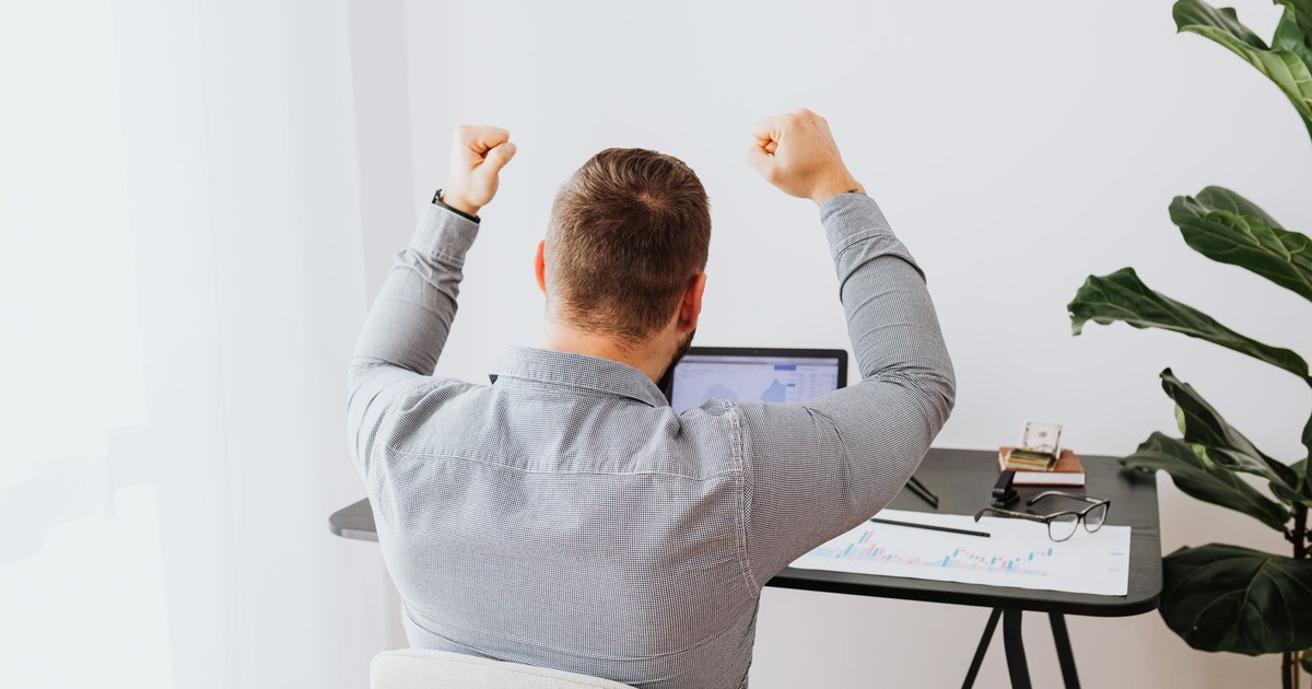 Back view of a man cheering with arms raised while sitting at a desk.