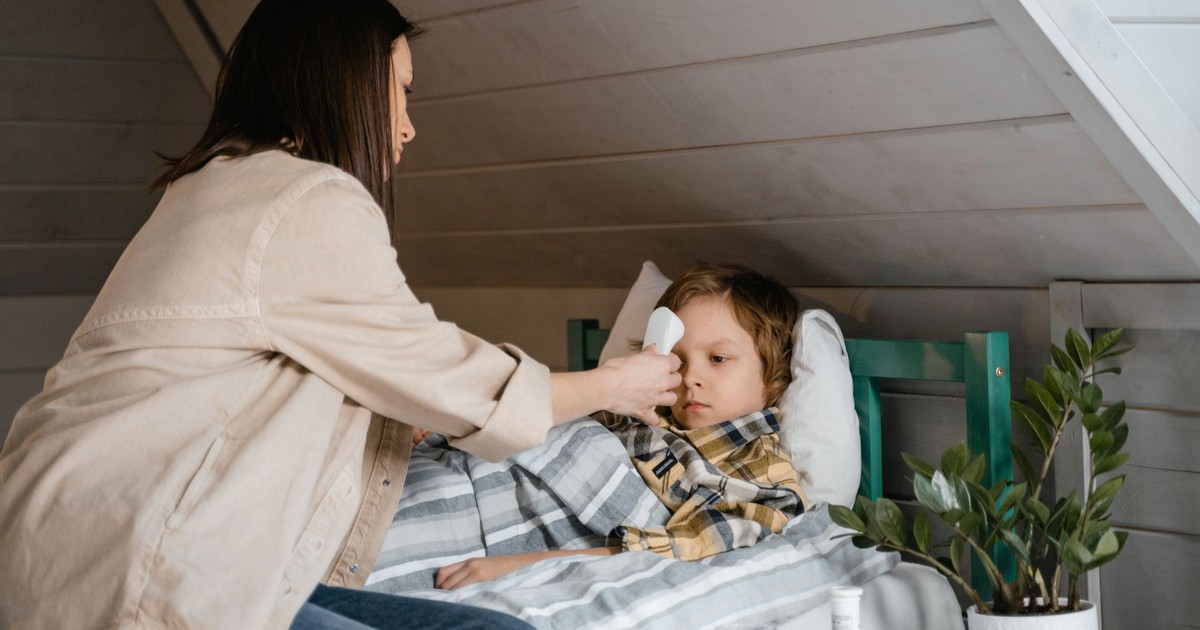 A mother checks her son's temperature, who lies sick in bed, inside a cozy bedroom.
