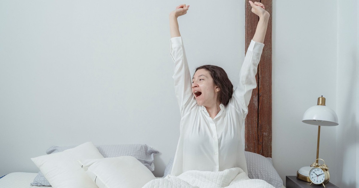 Sleepy brunette sitting on white comfortable bed under blanket and stretching arms up while yawning and looking away during morning time at home