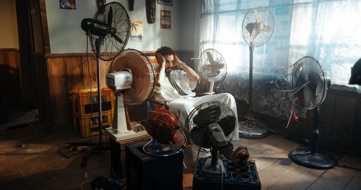Man in white outfit sitting surrounded by several fans indoors during daytime.