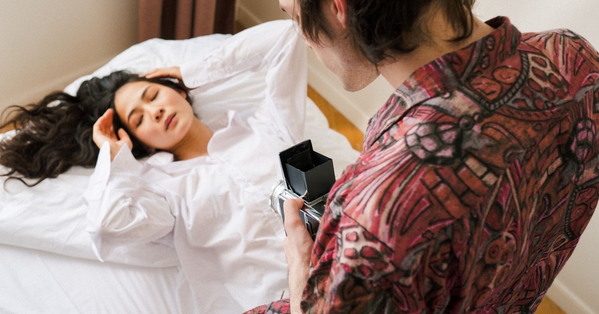 A man photographing a relaxed woman in a cozy bedroom setting.