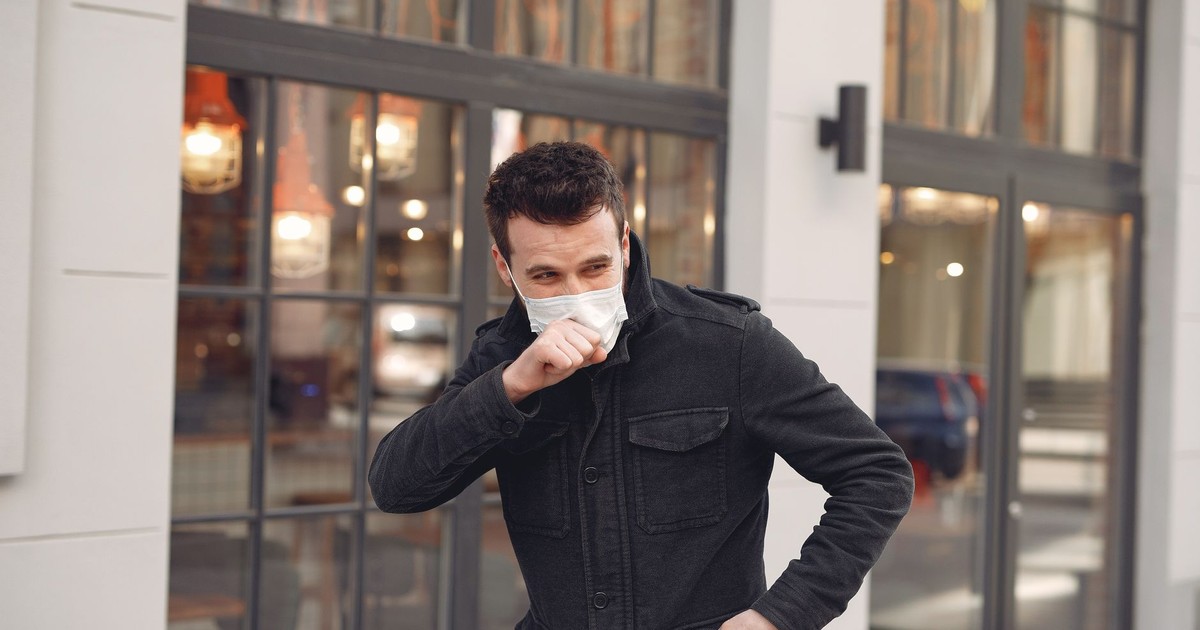 Unhealthy male wearing warm jacket and protective facial mask coughing while standing alone on urban street against city restaurant exterior in cold season
