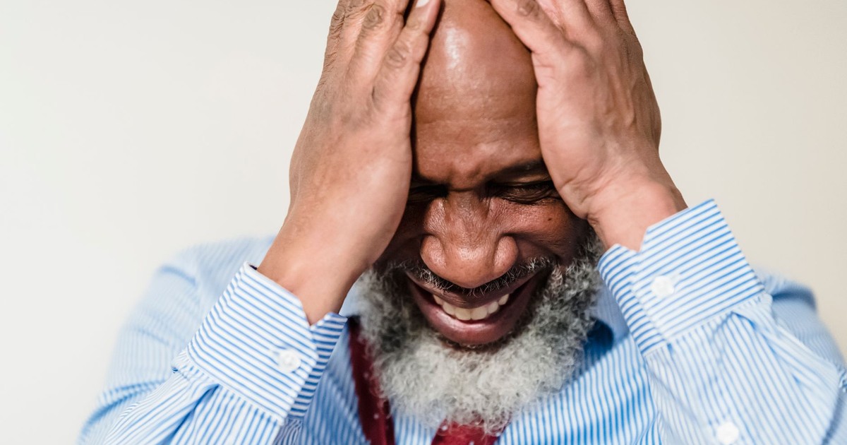 Elderly man in striped shirt showing stress and emotion with hands on head.