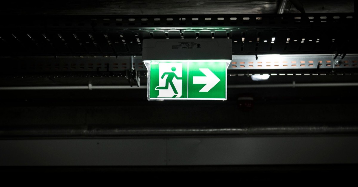 Bright green emergency exit sign with arrow in a dark industrial setting, indicating evacuation route.