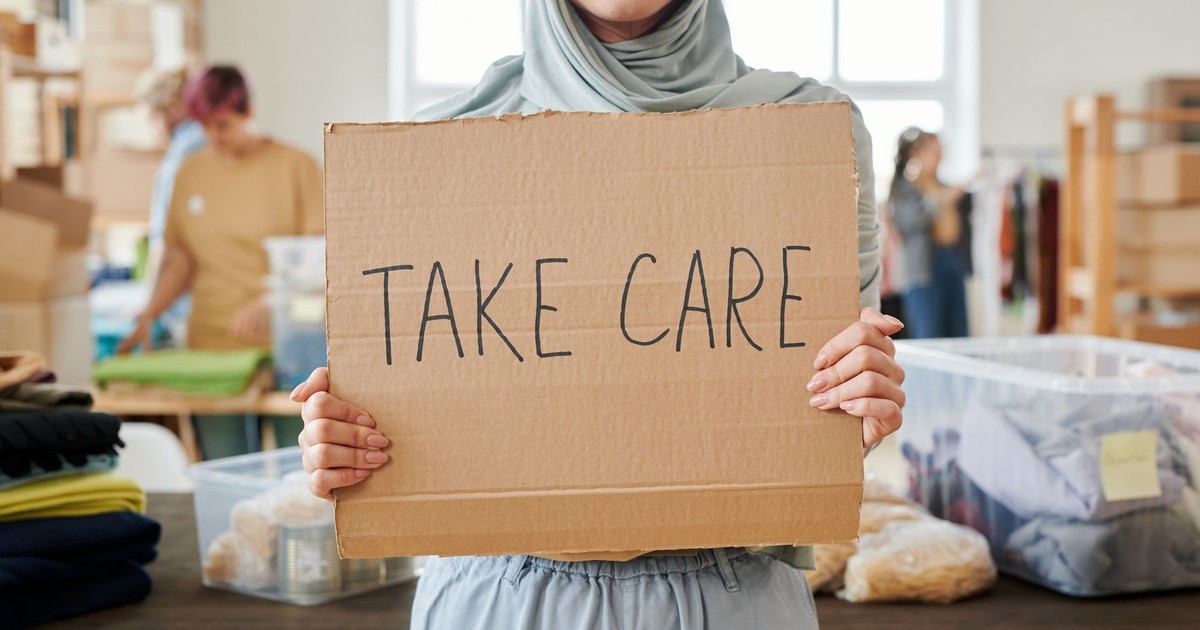 Smiling volunteer in hijab holding a 'Take Care' sign at a charity event indoors.
