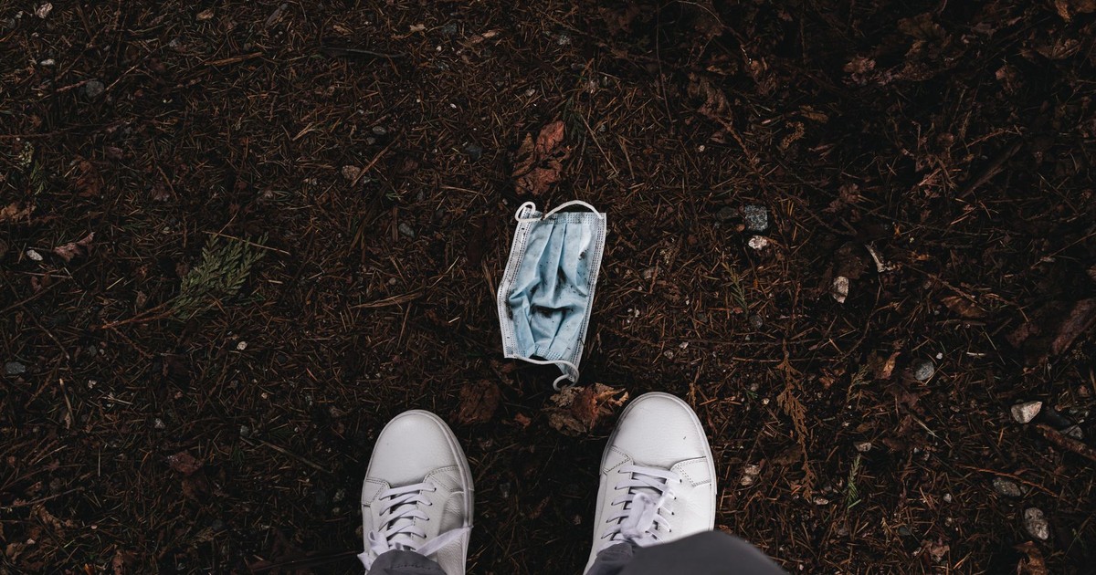 Top view of a discarded surgical mask on the ground surrounded by dried leaves, seen from above with white sneakers.