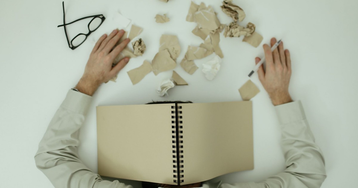 Overhead view of an exhausted man resting with a notepad over his head and crumpled papers around.