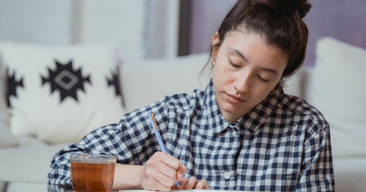 Young woman in checkered shirt writing in notebook with tea on glass table.
