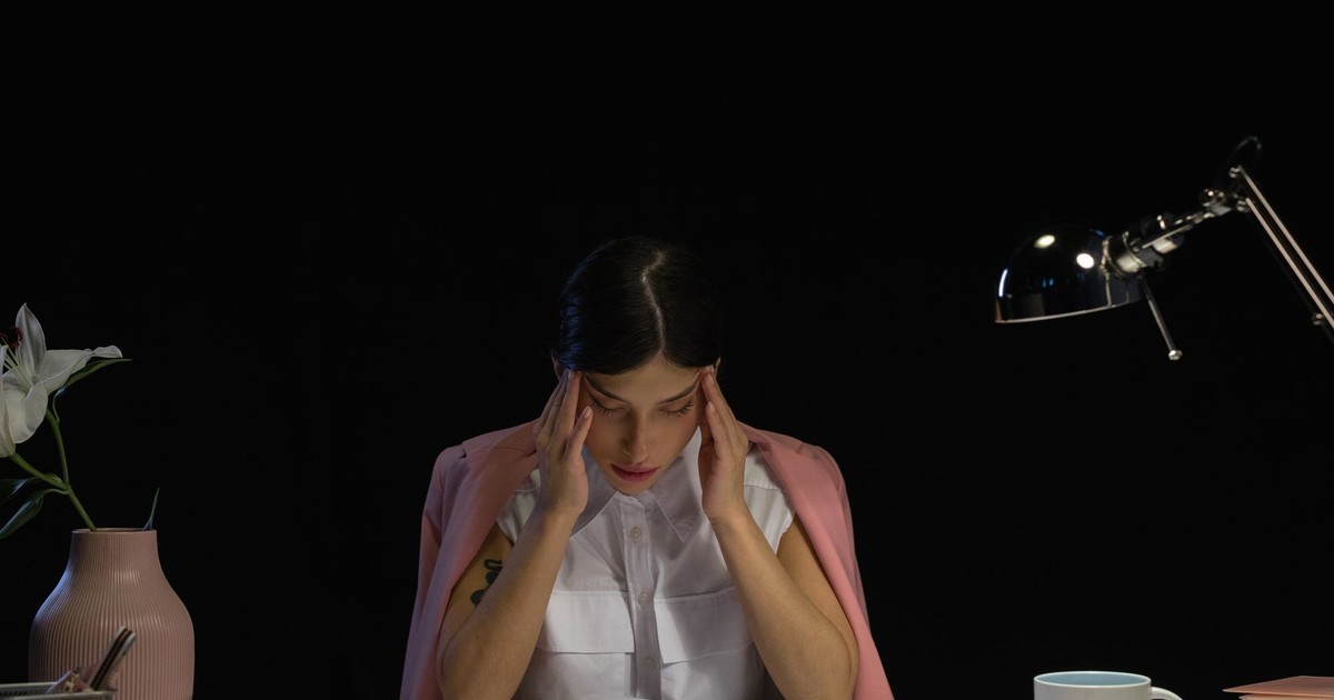 A woman sitting at a desk with her hands on her head, appearing stressed and tired with office supplies around her.
