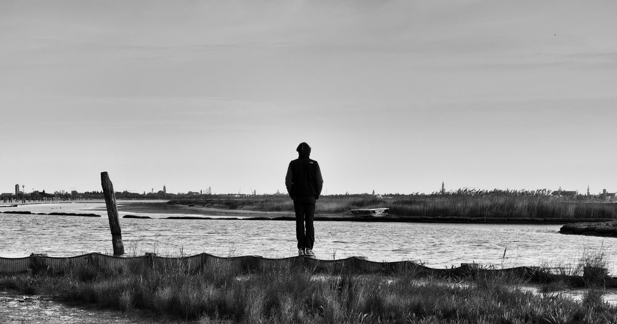 A person stands on a rural riverbank, embracing solitude and nature's serenity in black and white.