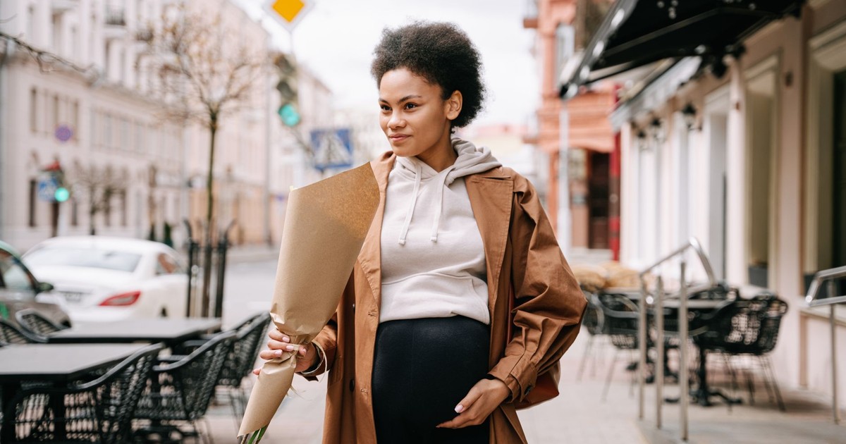 A stylish pregnant woman with afro hair holding flowers, walking down a charming city street.