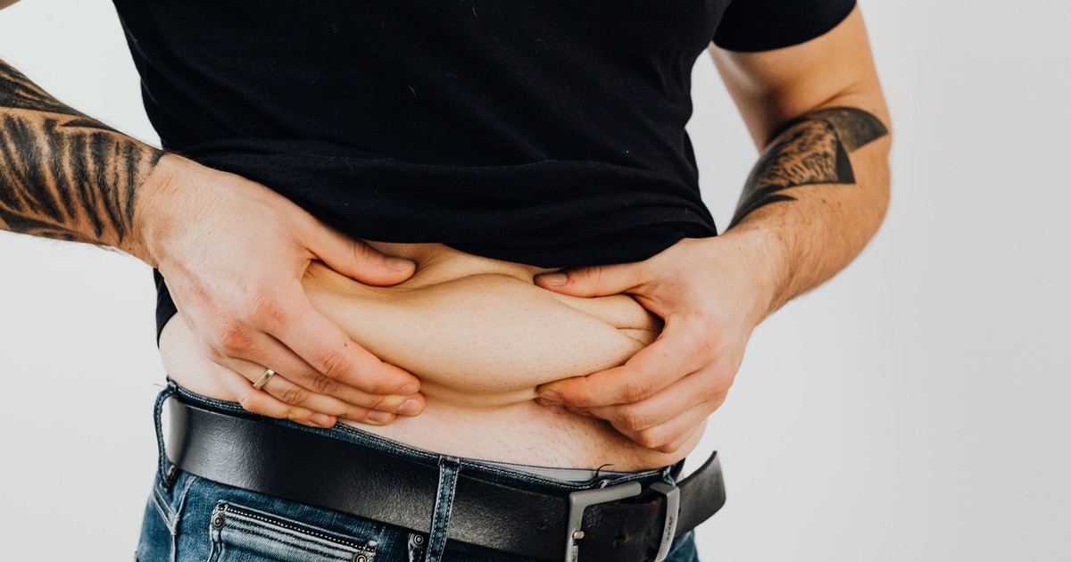 Close-up of a man pinching his stomach fold, wearing a black shirt and jeans with visible tattoos.