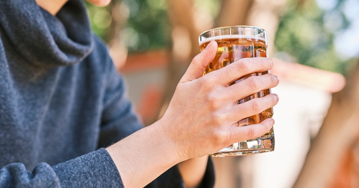 A person in a black jacket holding a clear glass of beer outdoors on a sunny day.