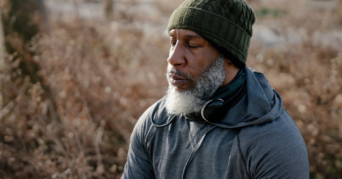Bearded senior male wearing warm clothes with headphones on neck sitting with closed eyes in nature