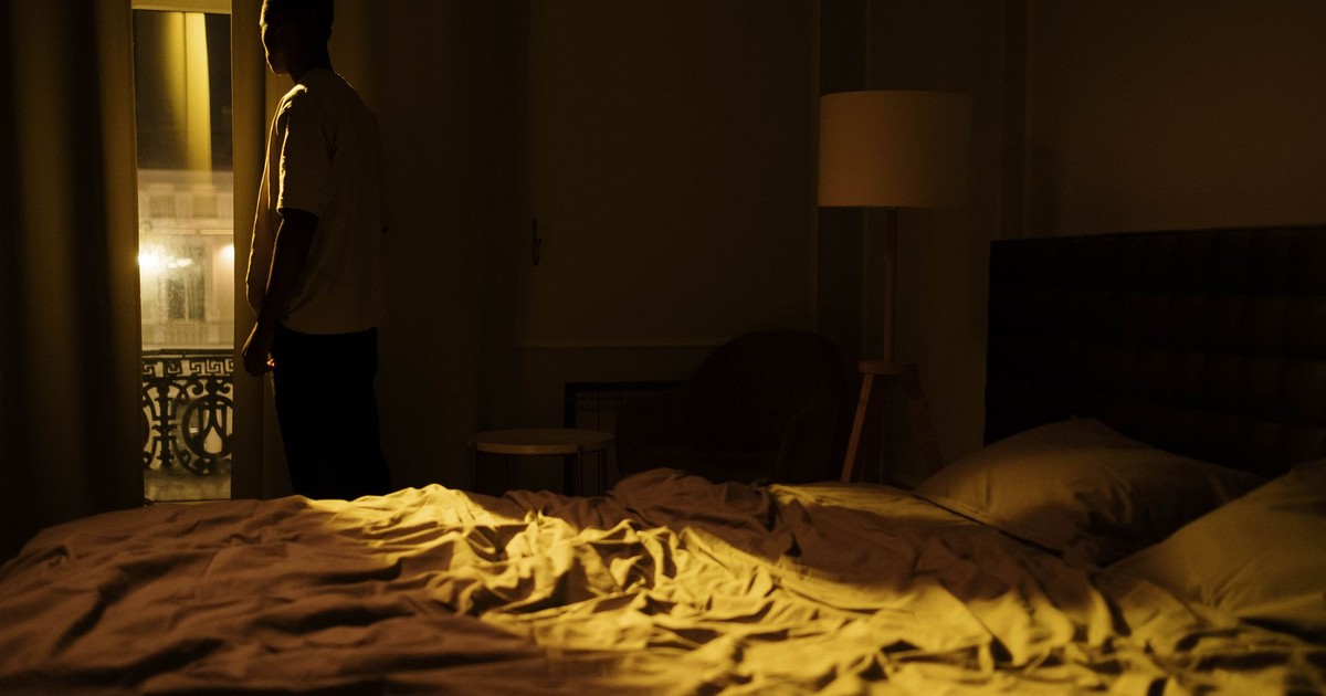 Man in dark bedroom gazing out window, highlighting themes of insomnia and solitude.