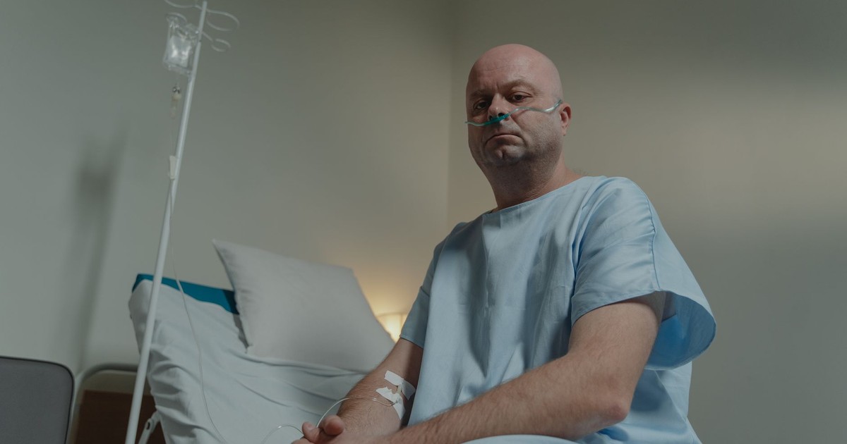 Bald man in hospital gown sitting on bed with nasal cannula and IV in a clinical setting.