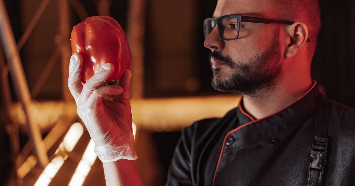 Chef examining a fresh bell pepper in a dimly lit kitchen setting, focusing on food quality.