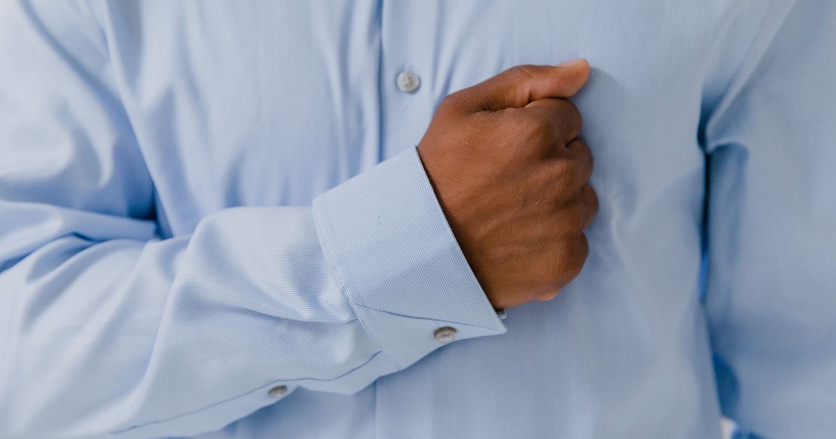 A close-up of a man's hand pressing on his chest, wearing a light blue shirt, showing a gesture.